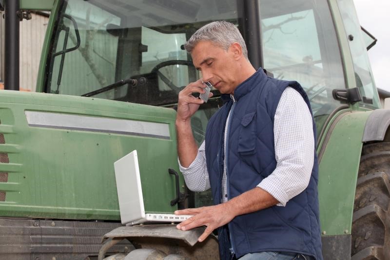 Famer looking at his laptop while on his phone. Tractor in background