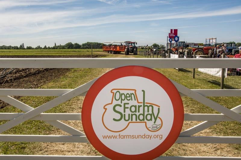 An image of the Open Farm Sunday sign on a farm gate, with a tractor and trailer in the background. 