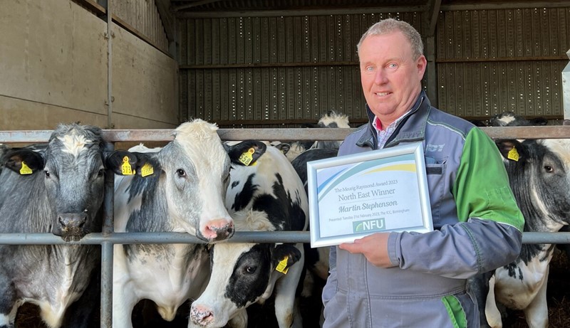 Martin Stephenson stands in front of his cattle shed, with cows looking out, holding his framed winner's certificate 