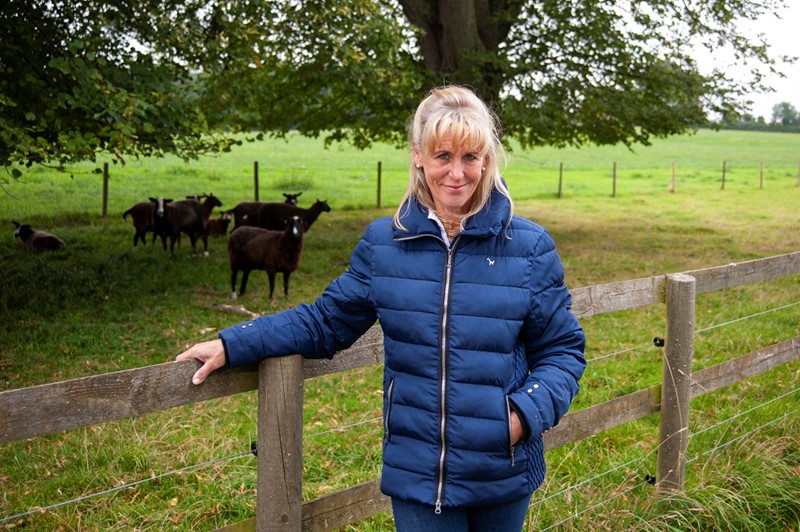 NFU President Minette Batters standing in a field, leaning against a wooden fence with sheep grazing behind her.