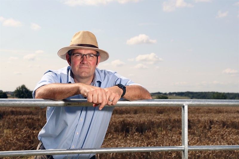 A picture of Tom Bradshaw wearing a hat, leaning on a gate to a field of crops