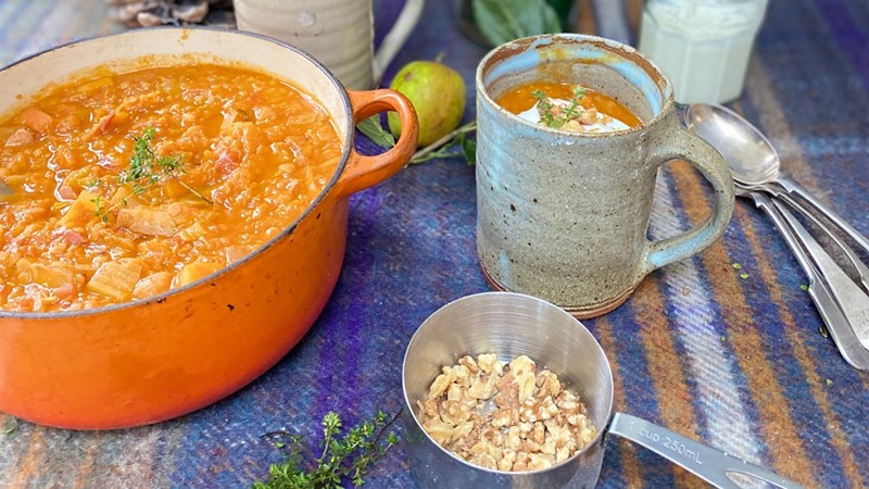 An image of lentil, bacon and roasted winter vegetable soup in an orange dish, with another portion in a ceramic mug and nuts in a small saucepan. 