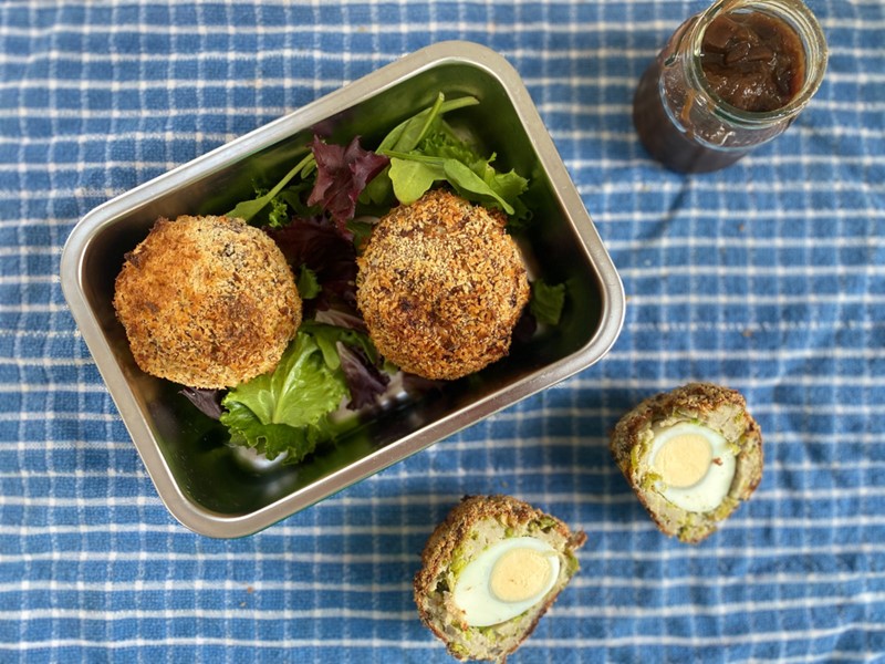 Two golden Scotch eggs, served in a metal tin on a bed of salad leaves, placed on a blue checked table cloth