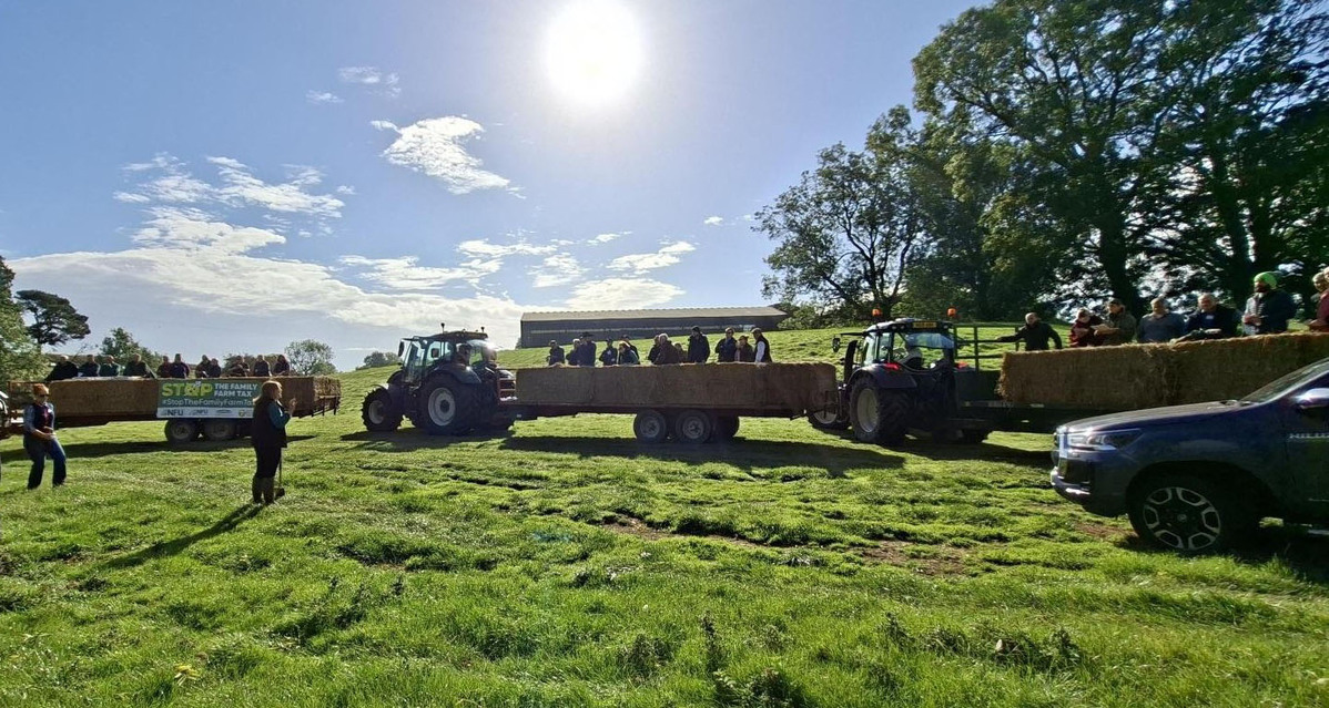 A photo of three tractors filled with people in a field listening to a speaker with the sun shining above