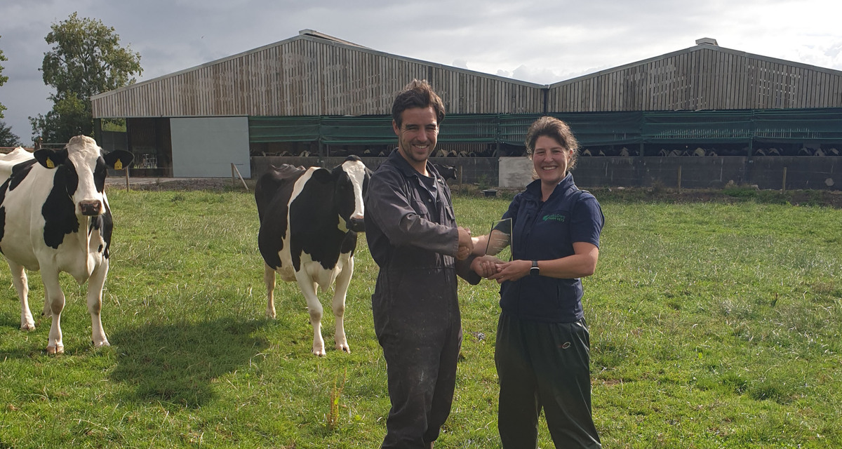 Vet Isobel Johnston handing Cheshire farmer Rob Yarwood with an award stood in a field with cows in front of a barn