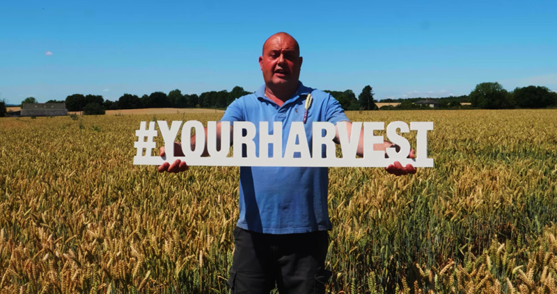 A picture of a farmer in a field, holding a sign that says #YourHarvest