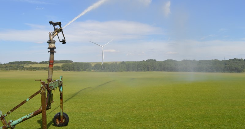 A photo of an irrigation system running in a field. A wind turbine can be seen in the background.