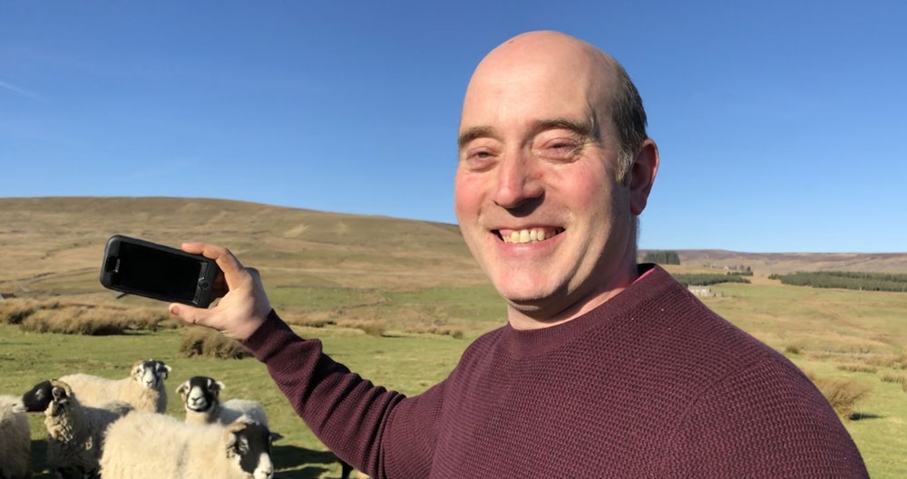 Greg Dalton on his farm with sheep and hills in the background on his phone doing FaceTime a Farmer