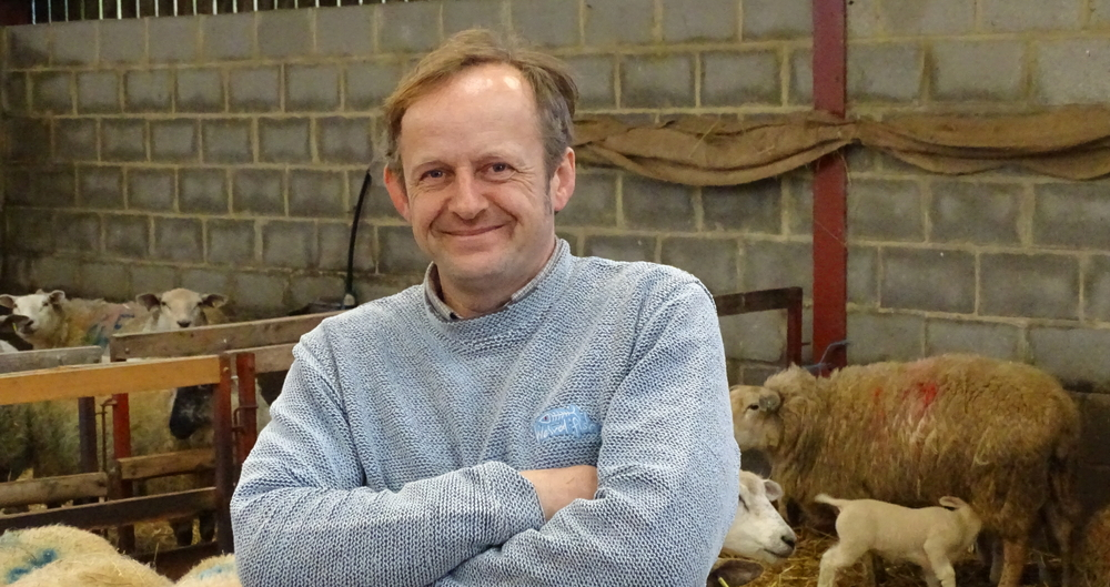 Will Terry smiling onto the camera, standing in his sheep shed with sheep and lambs behind